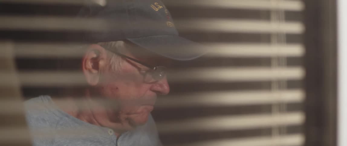 Elderly person sitting indoors wearing a hat and glasses