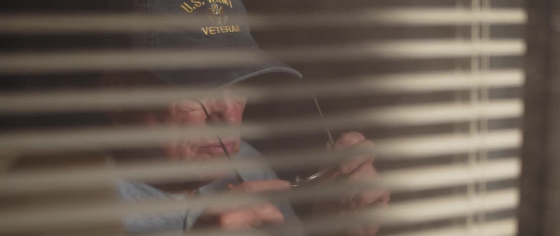 Portrait of an elderly person sitting and putting on their glasses while wearing a cap