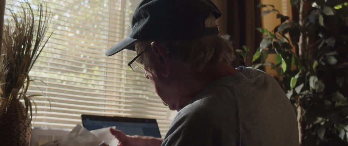 Elderly man wearing a cap glasses and gray t shirt works on laptop in room