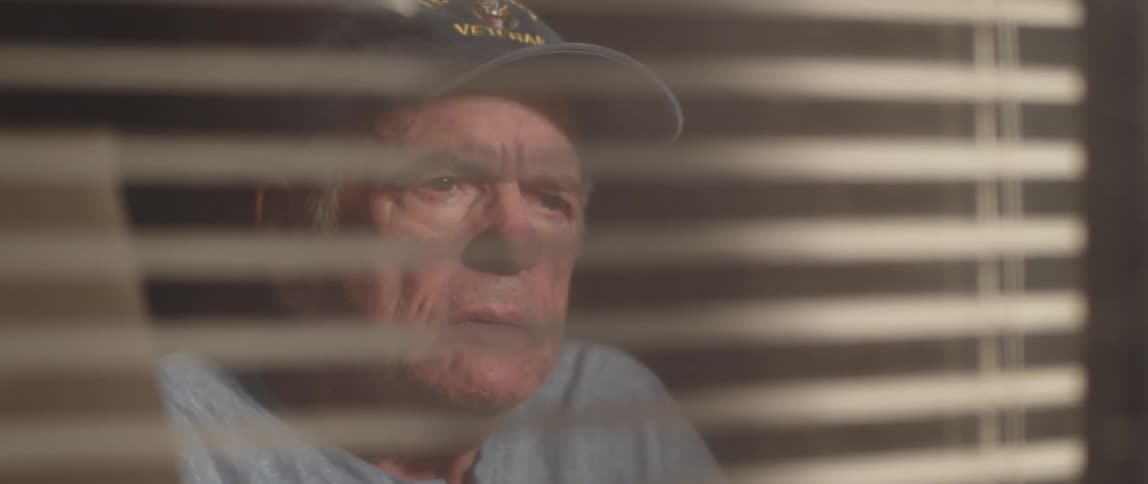 An elderly man wearing a cap looks through window blinds lost in contemplation