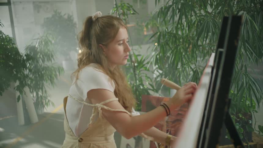 Woman sits painting in room full of scattered plants