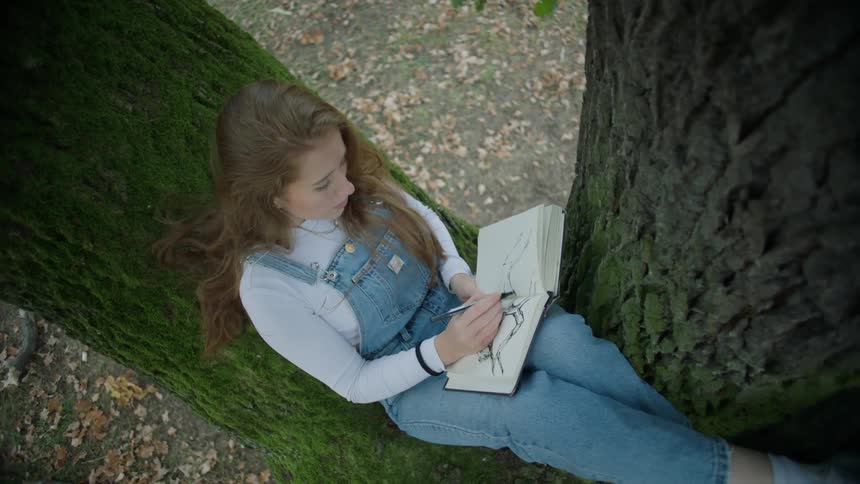 A young lady sitting on a tree drawing on her sketch book