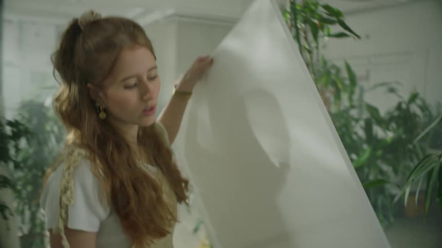 A woman walks across a room full of house plants to pick up a blank canvas which she puts on a stand before painting on it
