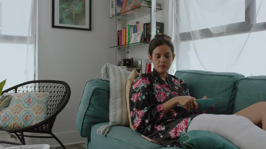 Woman with one leg bandaged sitting on the sofa eating and then picking up her phone feeling bored
