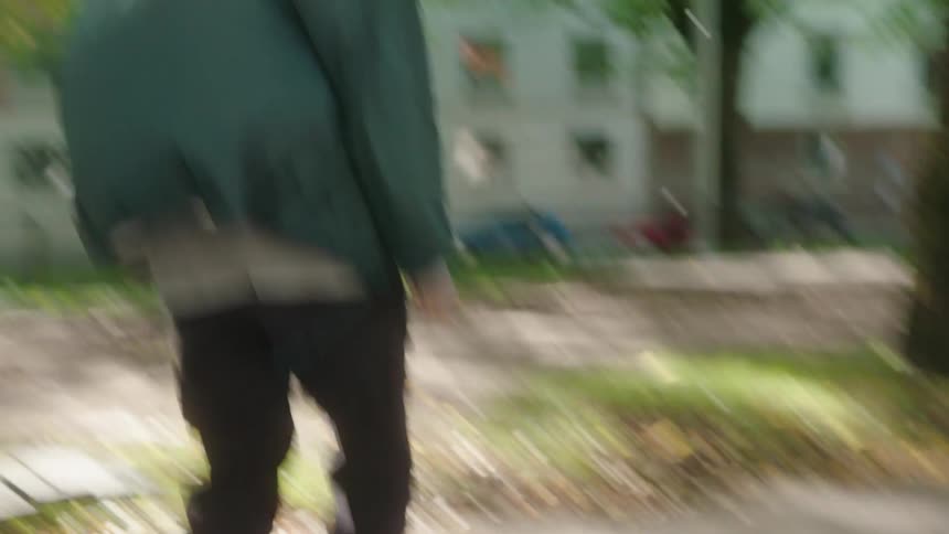 A long haired teenage boy is riding his skateboard on a neighborhood sidewalk and another skateboarder is behind him