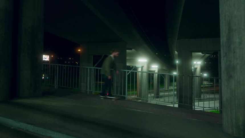 Men skateboard down road under bridge at night