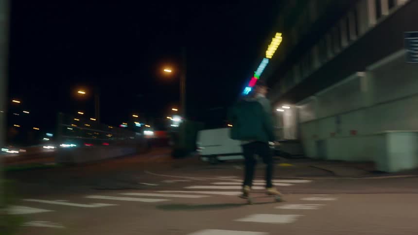 A pair of skateboarders skate along a road through a city at night