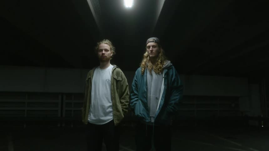 Two young men standing side by side in a parking garage with a skateboard at their feet
