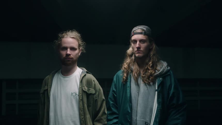A pair of men gaze into the camera while standing side by side in a dimly lit interior