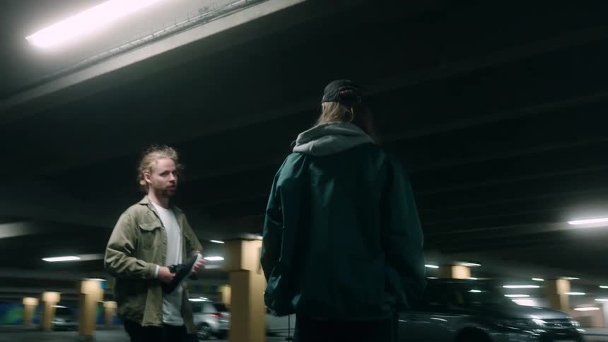 Two men on skateboards stand in a parking garage and talk