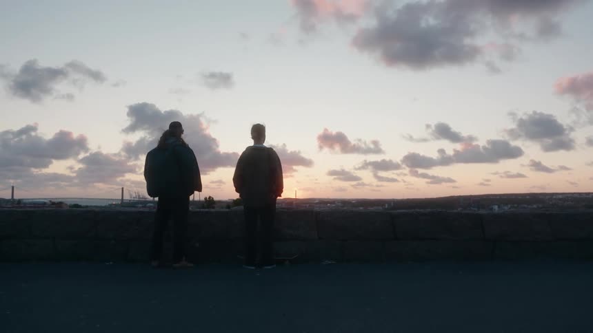 Two people stand near low wall looking toward horizon as setting sun turns sky red