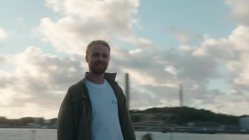 Young happy man stands with a skateboard on his hand on a park near a river at sunset