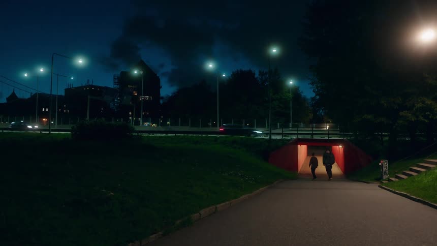 Two teenagers ride skateboards down a hill and through an underpass as traffic moves over the bridge above them