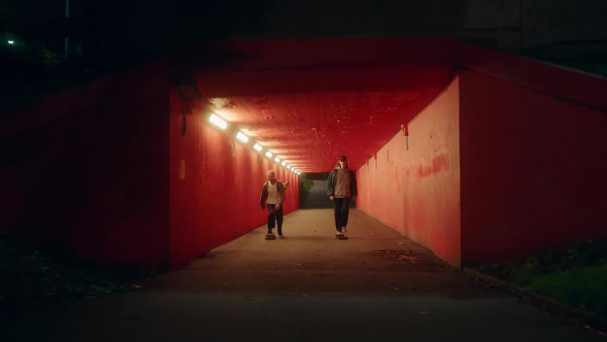Two skateboarders ride through tunnel together at night