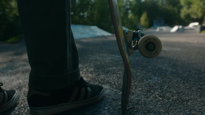 Sun shines brightly onto person standing in lot holding skateboard with ramp in the background