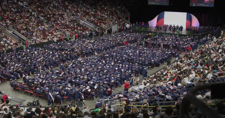 Panorama of a crowded grandstand with people in graduation gowns sitting on chairs on the court at a graduation ceremony