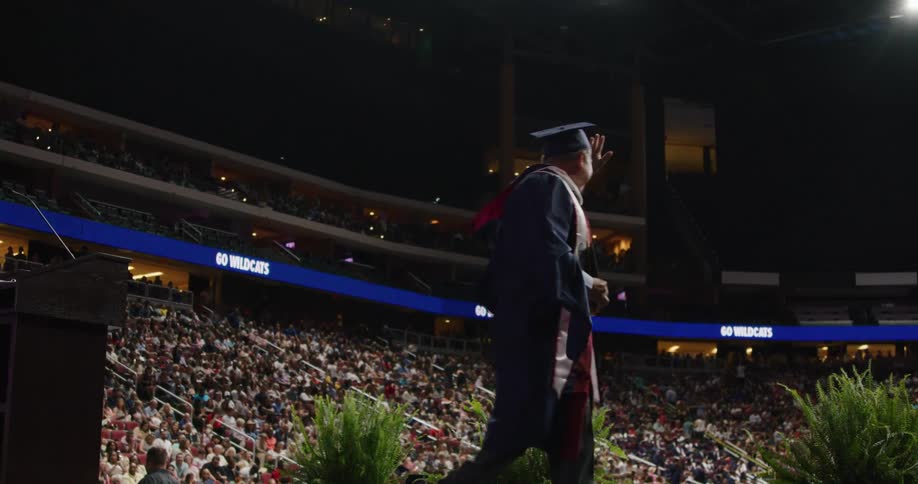 A male graduate walks across the stage and waves to the enormous crowd