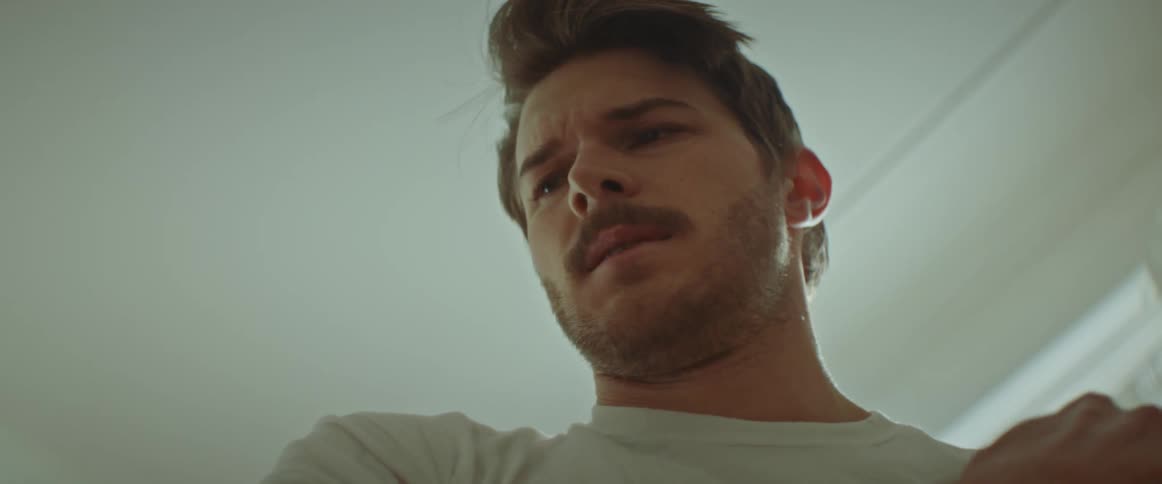 A man wearing a white t shirt stands indoors rotating shoulder and wincing in pain with the movement