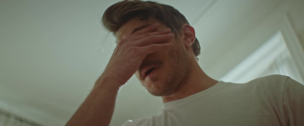 A man in a white t shirt leans his head back and repeating rubs his eyes with his hand