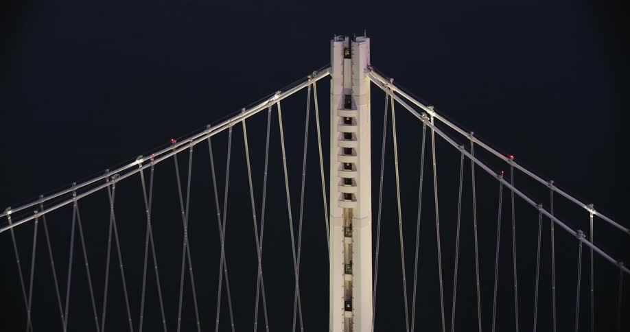 Aerial of the Bay Bridge Tower with cables extending out against the dark water below at night