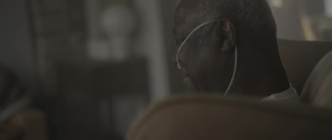 Elderly man with an oxygen tube sits smiling on a couch