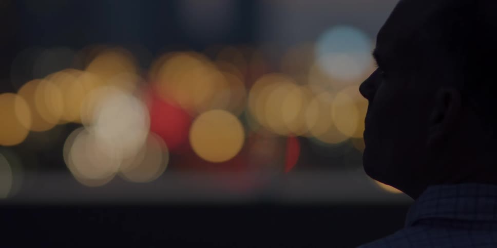 A man stares into the night as city lights illuminate