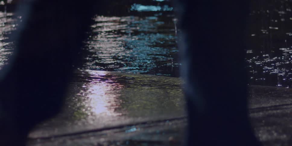 Rain falling on a city street at night with people walking on the sidewalk and cars driving down the street