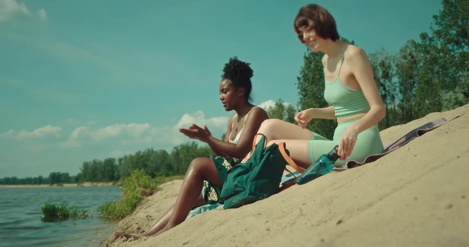 Two women sit on shore and smile while putting on sunscreen