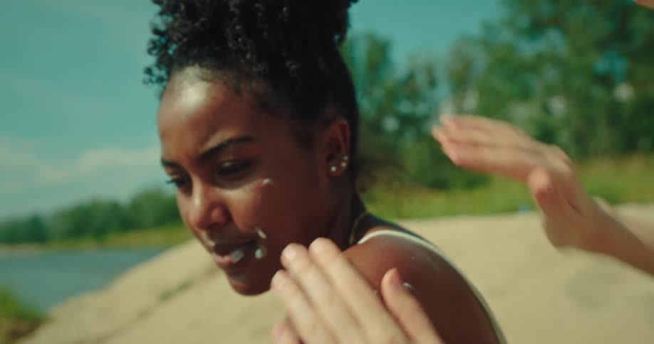 Friend applying sunscreen to her friends back and face at the beach
