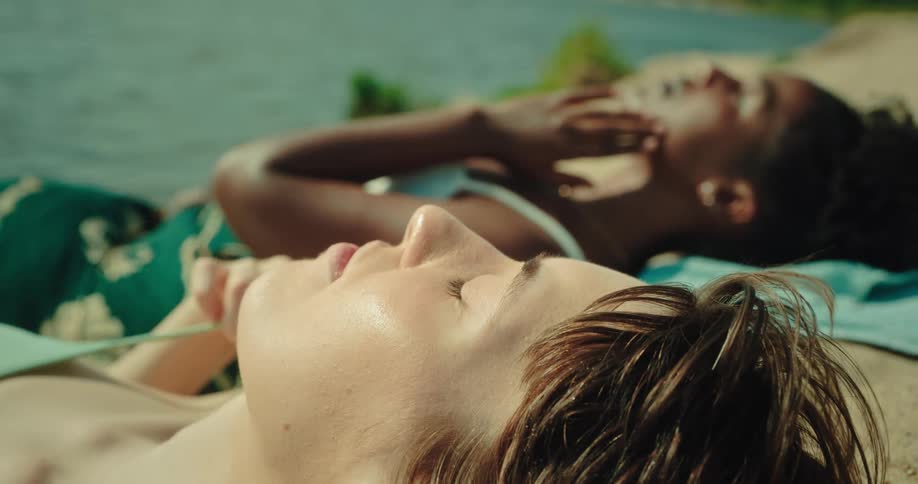 Two women are lying down with their eyes closed sunbathing next to the water of a sea