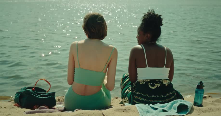 Two women touch their noses and then lie down to sunbathe in front of the sea