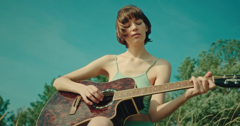 Female guitarist sits in field on a summer day and plays a guitar