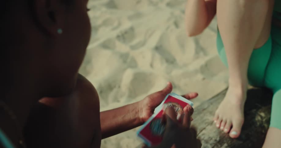 Two women playing a card game on top of a log on top of a sand