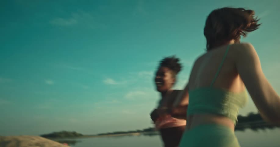 Two female friends spin around each other arm in arm on lake shore on a sunny day