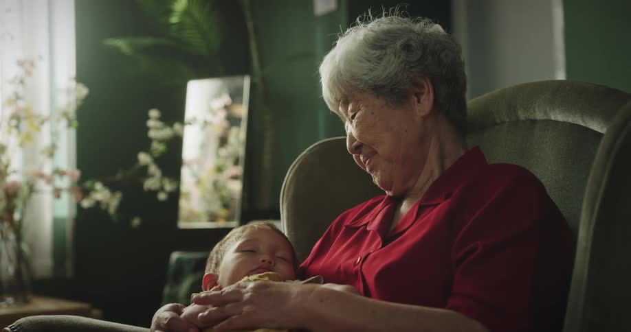 Grandmother smiles while sitting in chair and holding sleeping grandchild