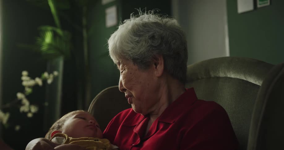 Woman placing the baby on her grandmothers lap who is sitting in an armchair