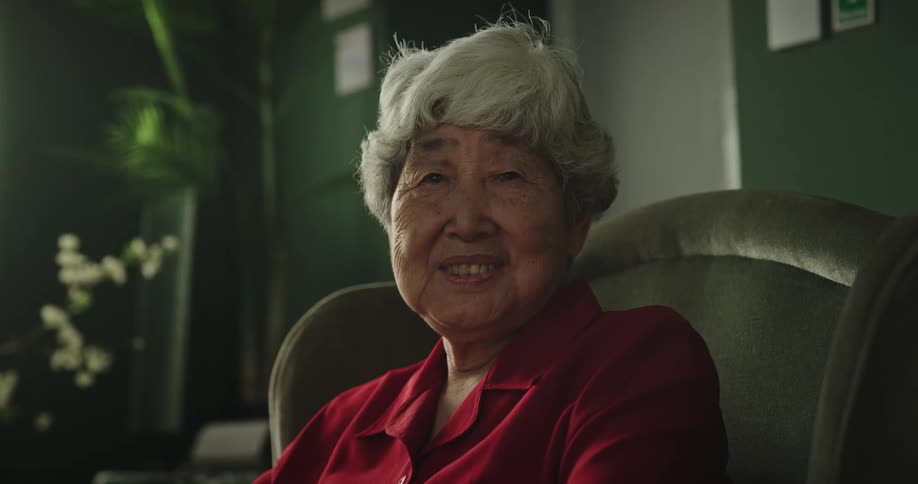 Elderly woman wearing a red shirt sits in a green armchair smiling