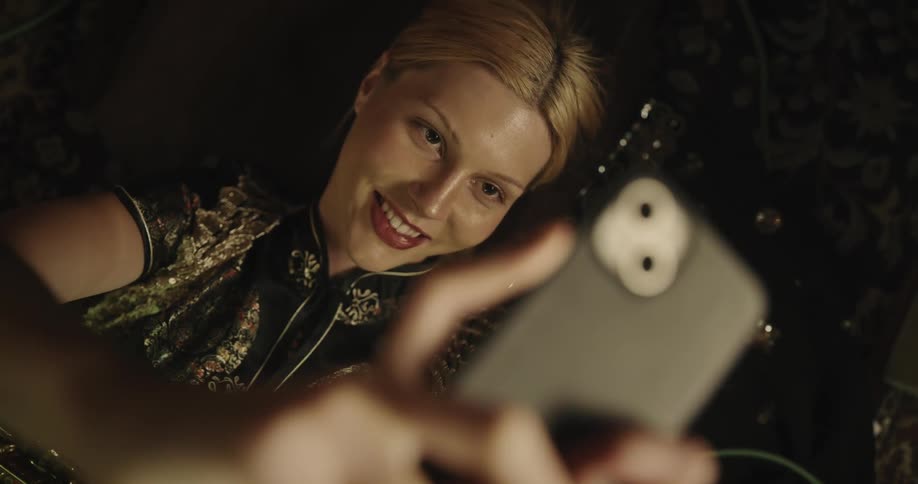 A woman is lying down with a saxophone on her lap and is pointing her cell phone at her face and making happy faces