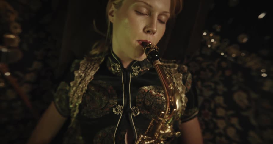 Woman leaning against pillow plays a saxophone without consulting nearby music sheet