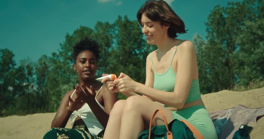 Women sit on beach smiling as they apply sunscreen