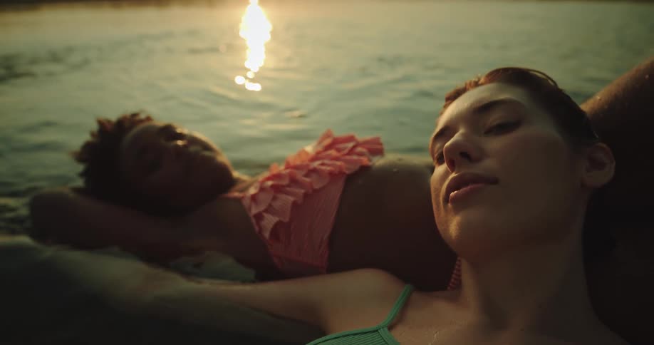 Two young women are lying in shallow waters relaxing one with her head on the others lap while the other caresses her head