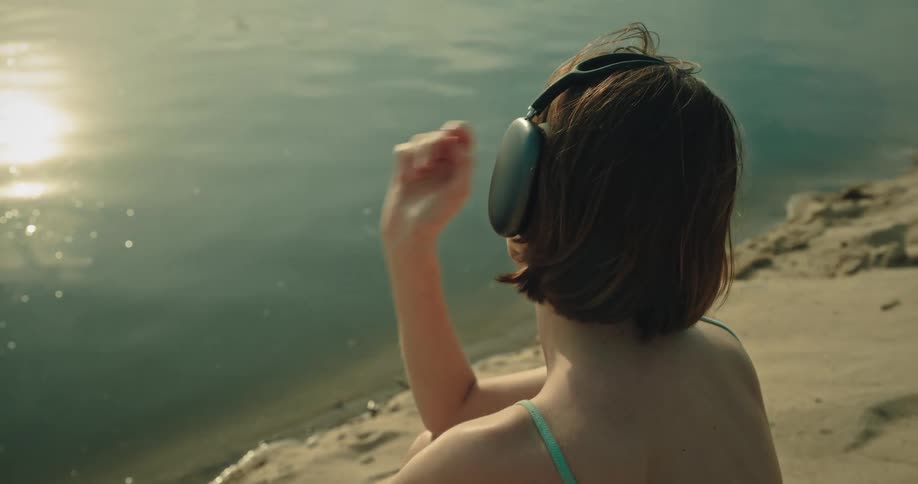 A female wearing headphones sits on the shore by the ocean