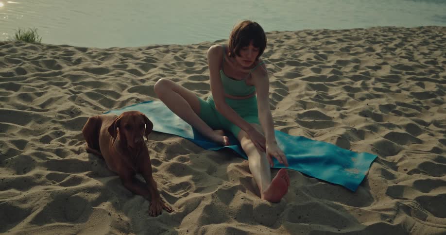 A young woman stretches while sitting on the beach next to her dog