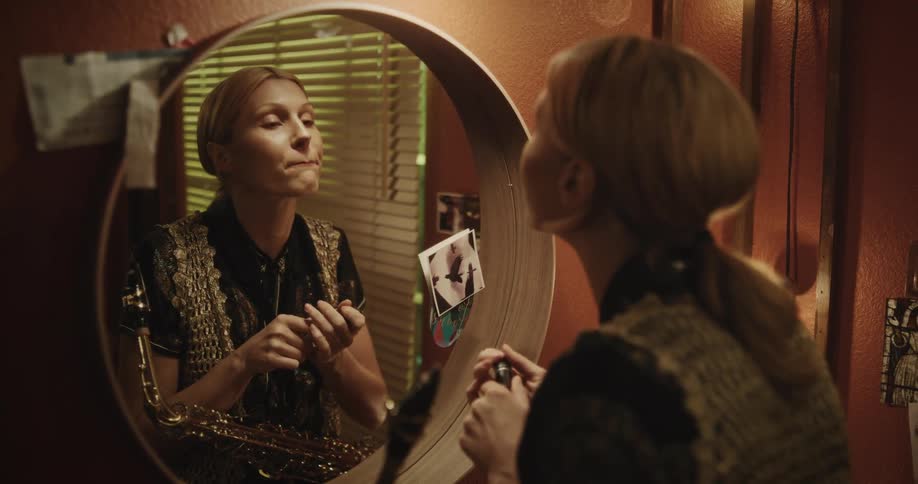 A woman puts on lipstick in the mirror and plays the saxophone