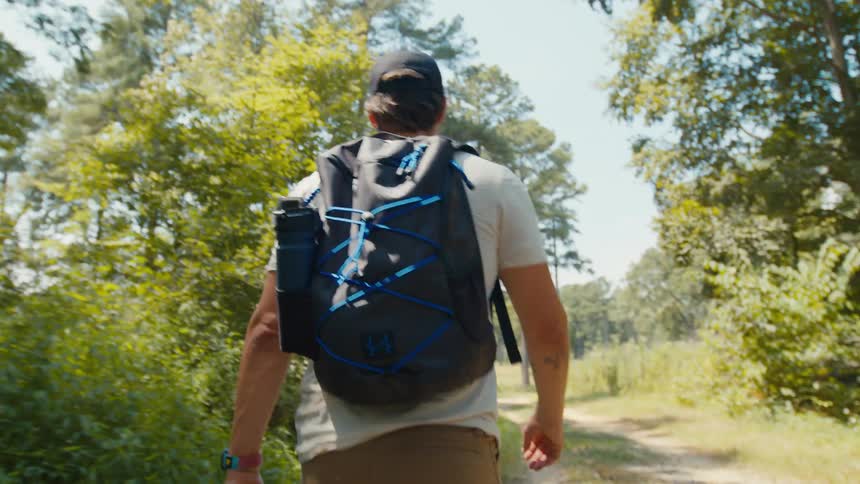 A man with a cap a white tshirt brown pants and a dark blue backpack walks along a dirt track in the forest