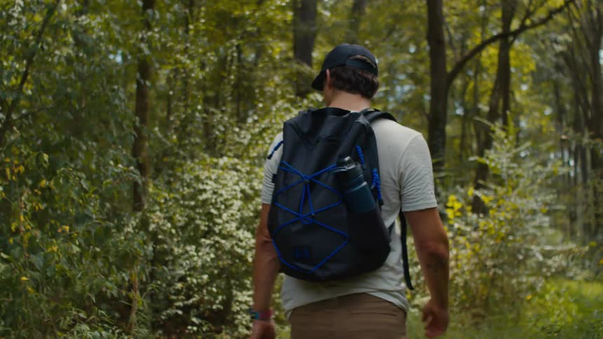 A man with a cap a white tshirt brown pants and a dark blue backpack walks along a dirt track in the forest