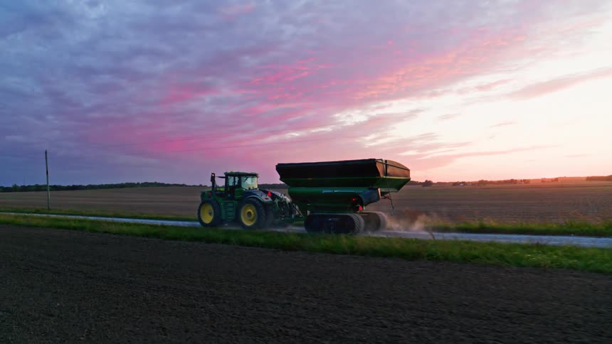 Tractor with harvest cart drives through beautiful Midwestern farmland at dusk with colorful pink and purple sunset
