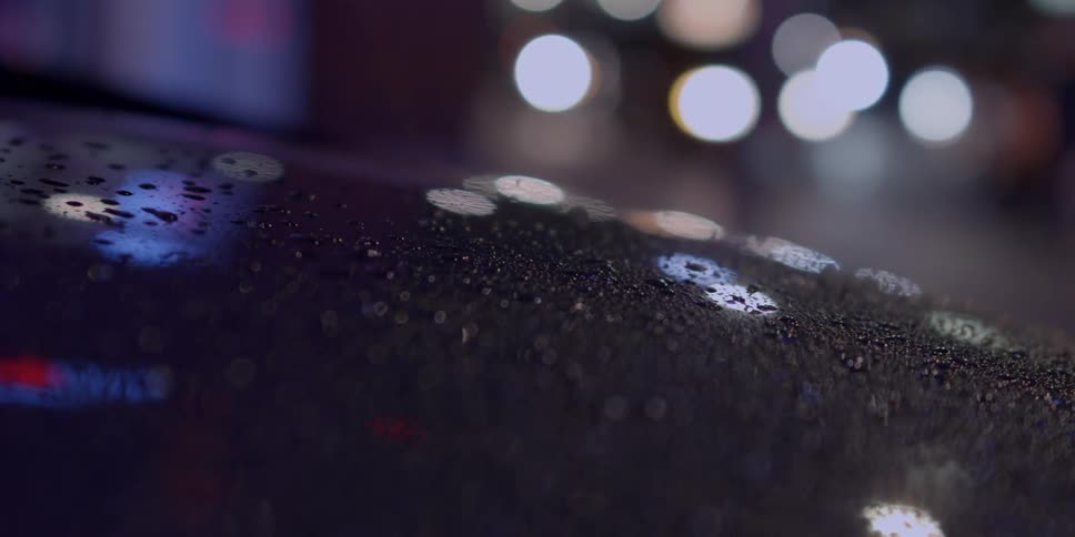 Water droplets sit atop a car as car lights illuminate the night