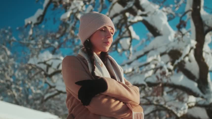 A teenage girl stands under a snow covered tree with her arms folded as she fixes her hat