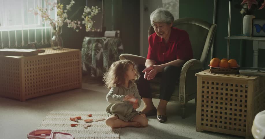 Grandmother sitting in the armchair with her granddaughter playing on the floor